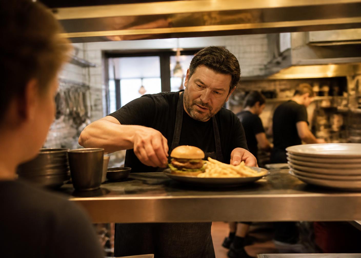 A chef plating a burger and fries at the kitchen pass of a busy neighborhood restaurant